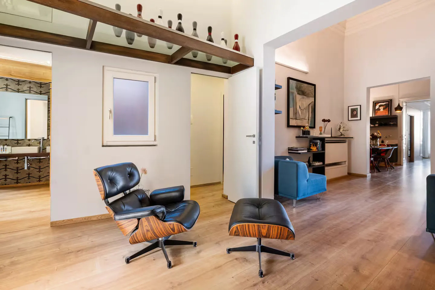 Il living visto dalla lobby: poltrona Eames in palissandro e nero su pavimento in rovere, poltroncina cobalto sul fondo, birilli da bowling allineati su una trave in legno
