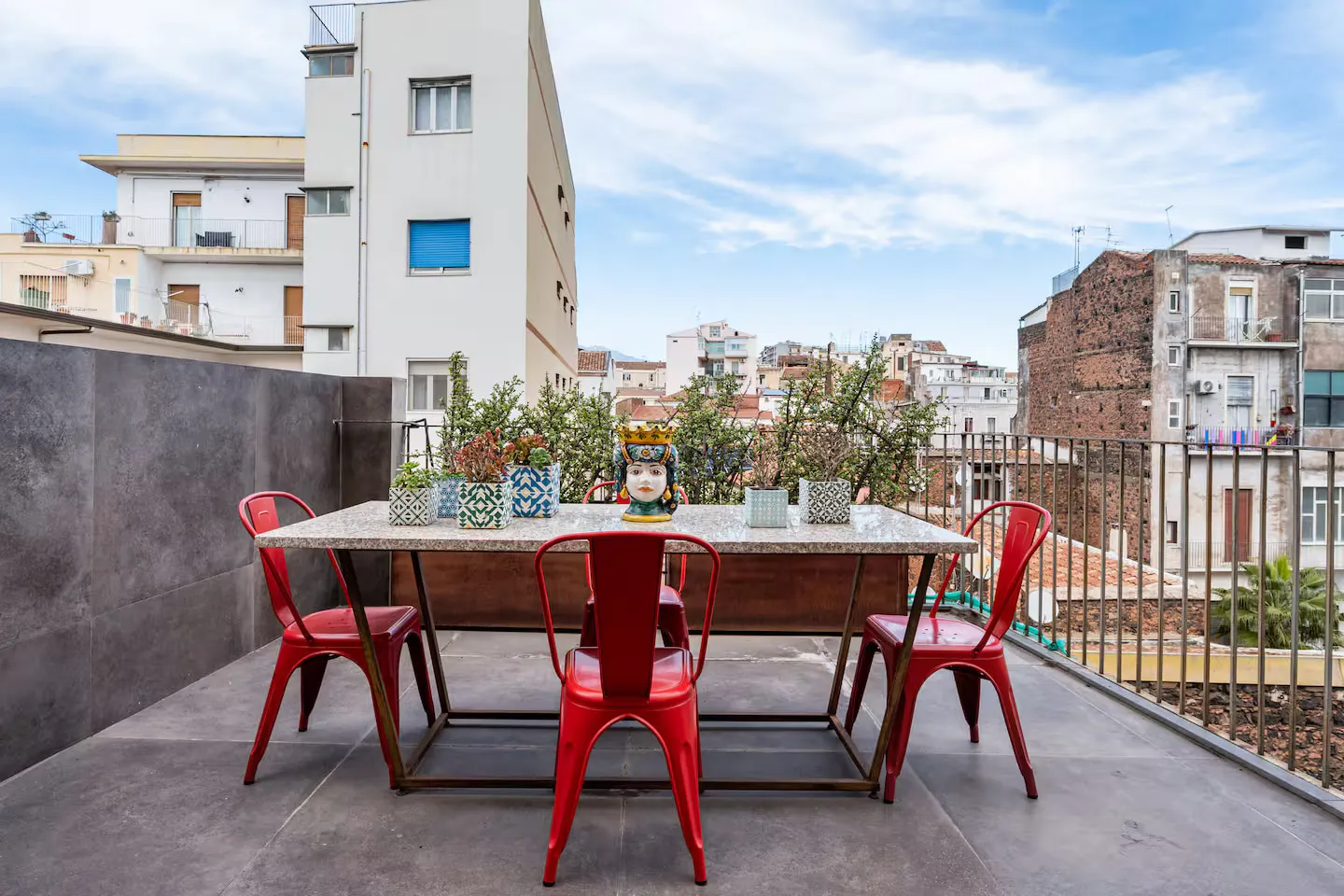 Il patio: tavolo con piano in marmo e base in ferro, quattro sedie Tolix rosse, vasi in cementina e una testa di moro fra i tetti di Catania