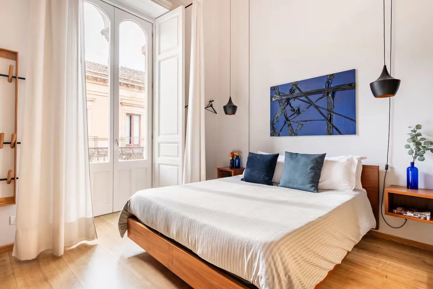 Room 02: letto con biancheria bianca e cuscini blu cobalto, quadro astratto blu alla parete, porte-finestra ad arco aperte sul balcone con tende leggere