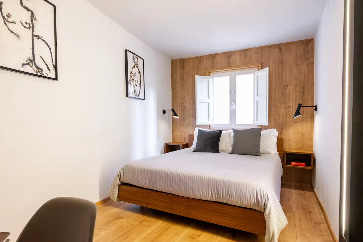 Room 03: letto addossato a una parete rivestita in legno, con finestra a battenti bianchi al centro e applique nere ai lati